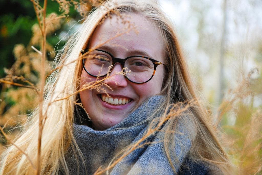 Man sieht Carlotta, wie sie in die Kamera lächelt und vor einem Auge eine Schilfblüte ist
